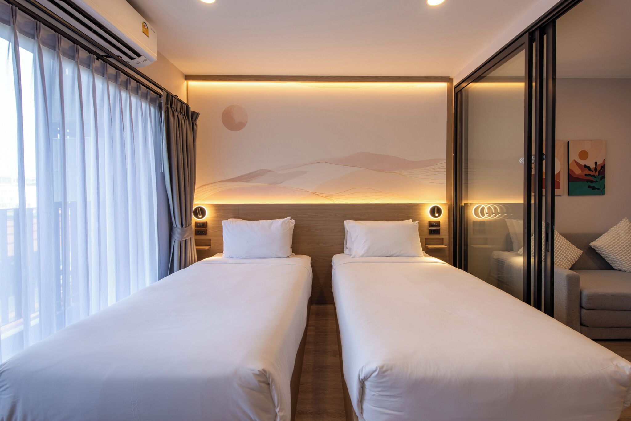 Une chambre d’hôtel avec deux lits, une grande fenêtre avec des rideaux, un climatiseur fixé au mur et un tableau représentant un paysage.