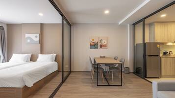 A modern hotel room with a bed, a dining table with chairs, a kitchen area, and artwork on the walls.