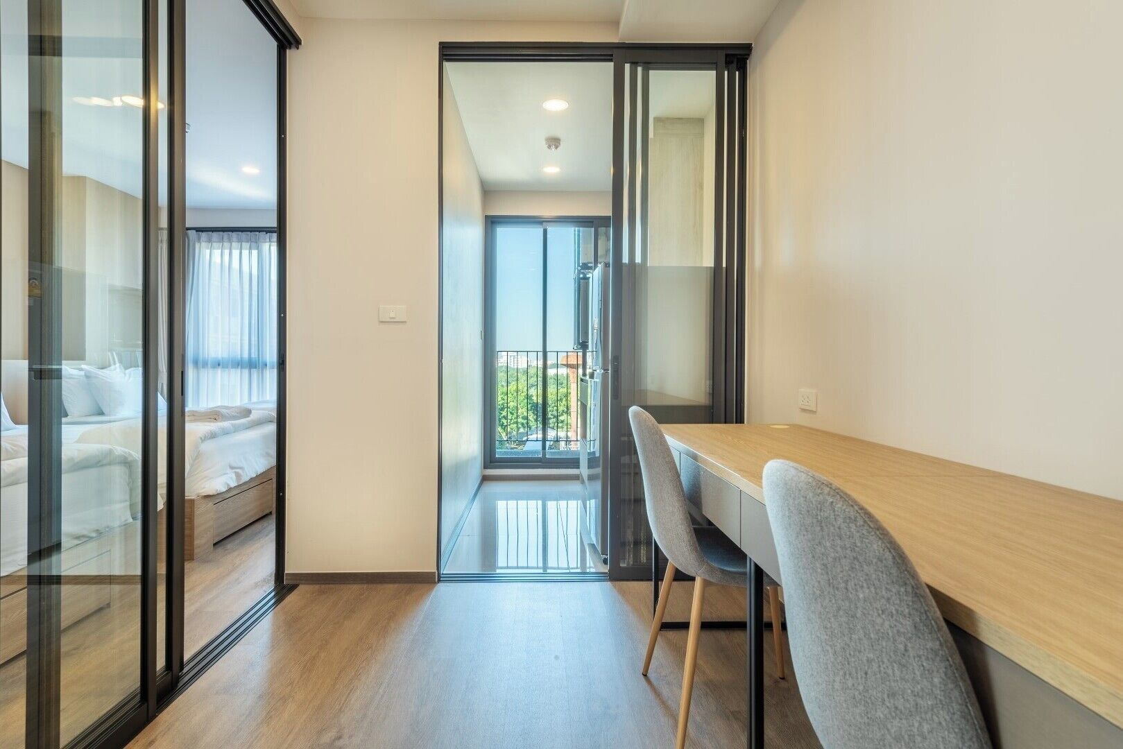A modern hotel room with a wooden floor, a glass partition separating the bedroom from the living area, a balcony with a view, and a dining table with chairs.