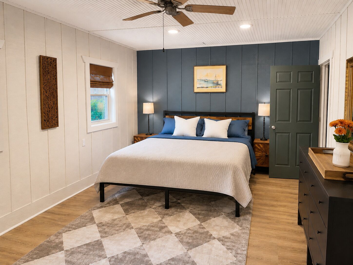 A bedroom with a bed, two bedside tables, a ceiling fan, a window with blinds, a door, and a dresser.