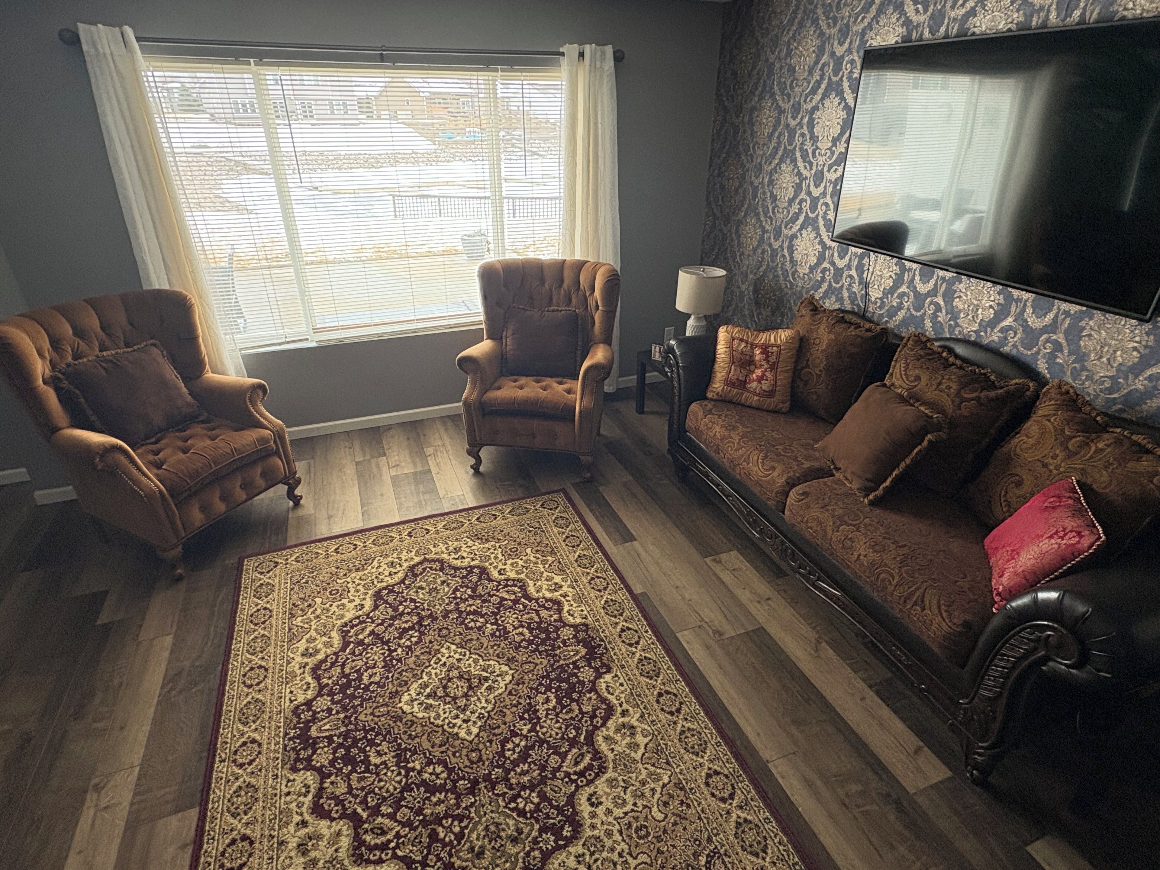 Een woonkamer met een groot raam, twee fauteuils, een bank, een vloerkleed met patroon, en een televisie aan de muur.