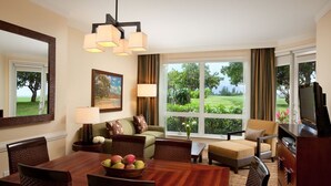 Habitación de hotel con comedor, sofá, televisión y ventanas amplias con vistas a zonas verdes.