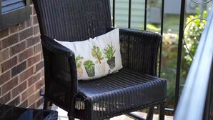 Une chaise en osier noir, dotée d’un coussin orné de motifs végétaux, est placée sur un balcon qui présente un mur en briques et une rambarde en métal.