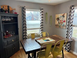 A dining area with a wooden table and yellow chairs, a dark cabinet with glassware, and a painting on the wall. - 3-bedroom House in Rapid City for Sturgis Rally with Two Car Garage for Bikes (Rapid City)