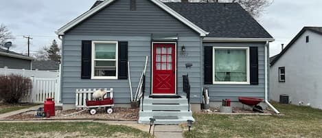 Une maison à deux étages avec une porte d’entrée rouge, une boîte aux lettres et un petit jardin devant.