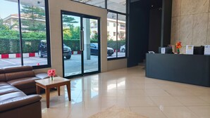 Een hotel lobby met een receptiebalie, een leren bank, een kleine tafel met een vaas, en grote ramen met uitzicht op geparkeerde auto's en groen buiten.