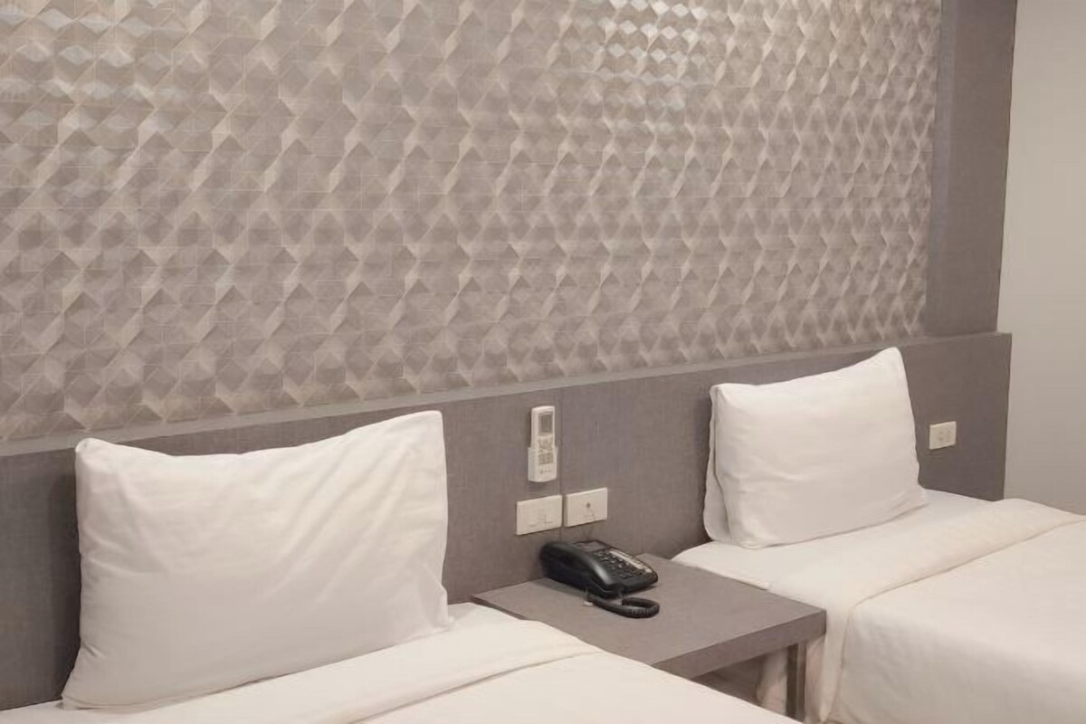 A hotel room with two beds, a textured wall, a bedside table with a phone, and a power outlet.
