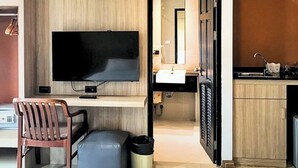 Een moderne kamer met een flatscreen televisie, een wand van houten panelen, een bureau met stoel, een klein voetenbankje, een prullenbak, en een kitchenette met spoelbak en kastjes.