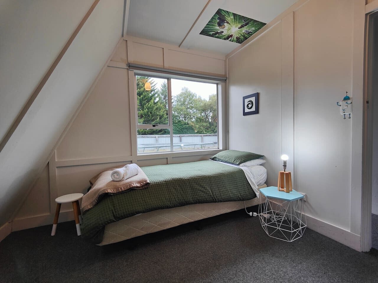 Ein kleines Schlafzimmer mit geneigtem Dach, einem Einzelbett, einem kleinen Hocker, einem Nachttisch mit Lampe und einem Fenster mit Blick auf Bäume.