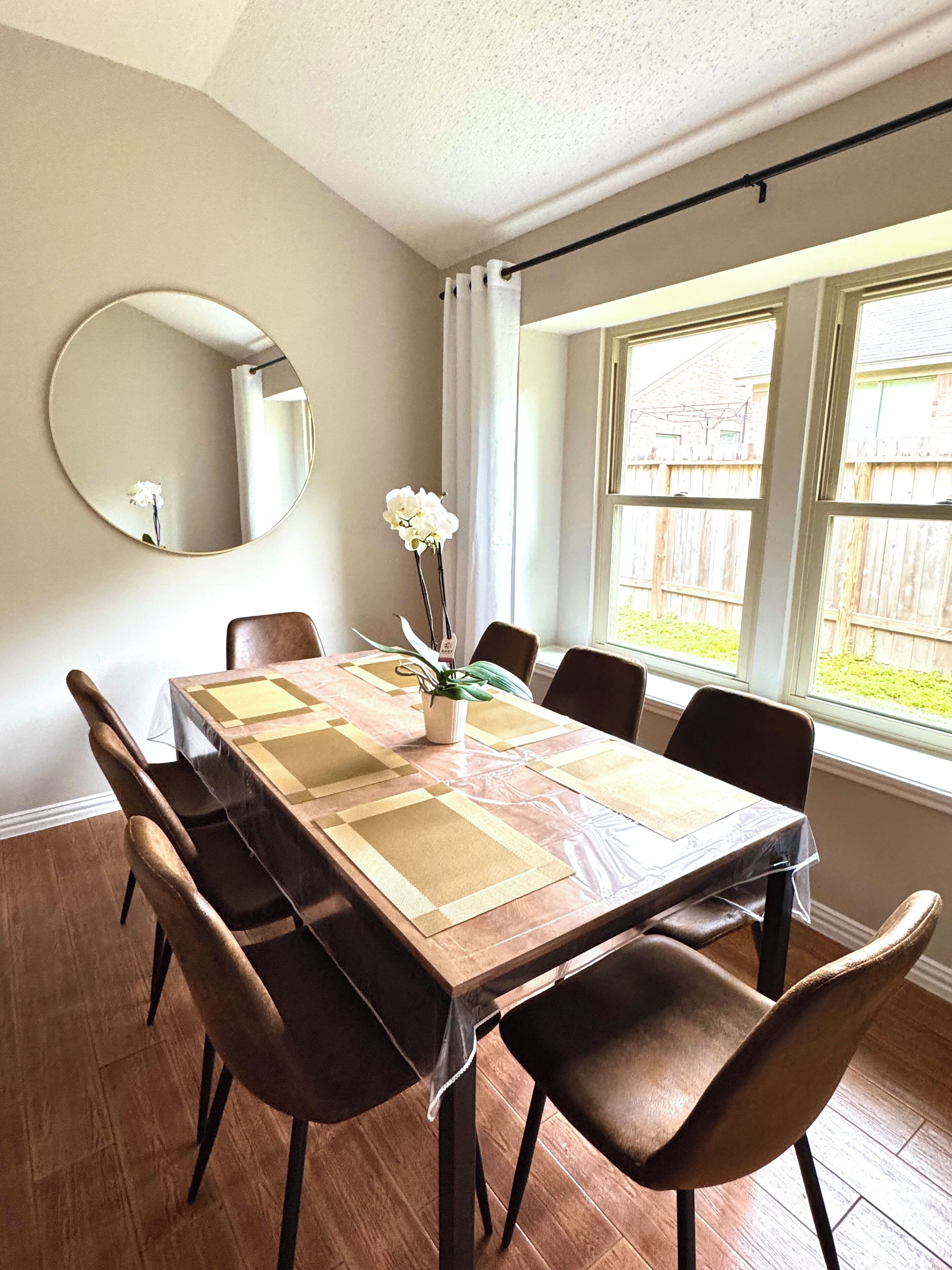Een eetkamer met een houten tafel, stoelen, een vaas met bloemen, een ronde spiegel en een raam met uitzicht op een houten schutting.