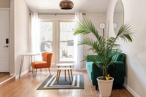 Una sala de estar con piso de madera, un sofá verde, un sillón naranja, una mesita blanca pequeña y una planta grande en maceta.