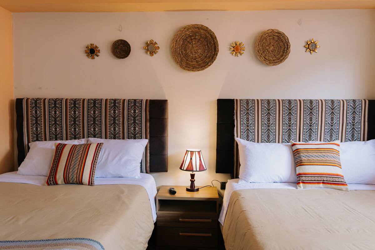 A hotel room with two beds, each with patterned bedding and pillows, a bedside table with a lamp, and decorative wall hangings.