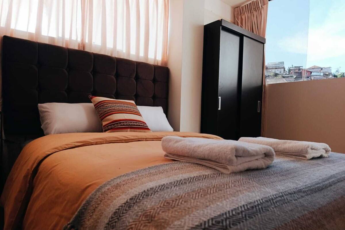 A neatly made bed with a patterned throw, a dark headboard, and a view of the city through a window.
