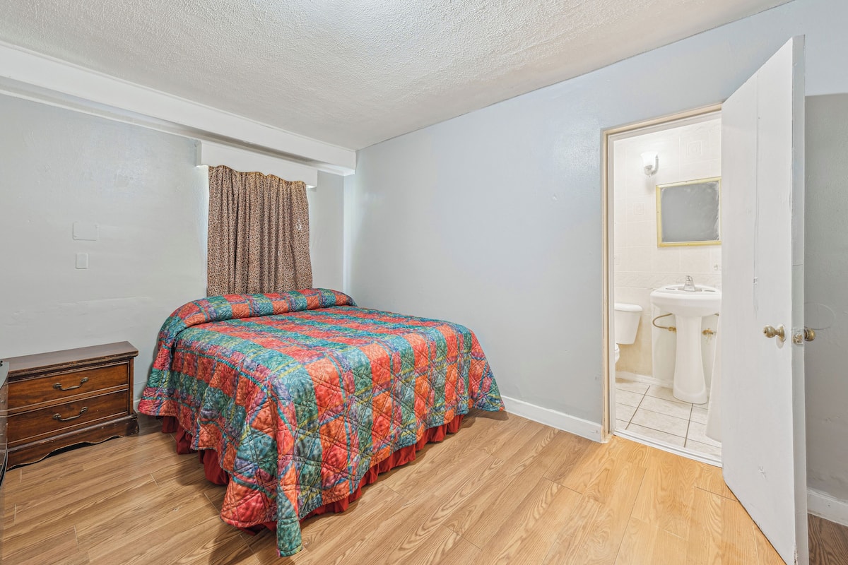A bedroom with a wooden floor, a bed with a colorful quilt, a wooden dresser, and a bathroom visible through an open door.