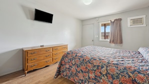 Een slaapkamer met een houten dressoir, een bed met bloemenbedekking, een flatscreen televisie aan de muur en een raam met gordijnen.