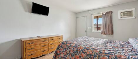 Un dormitorio con un aparador de madera, una cama con colcha estampada, un televisor de pantalla plana montado en la pared y una ventana con cortinas.