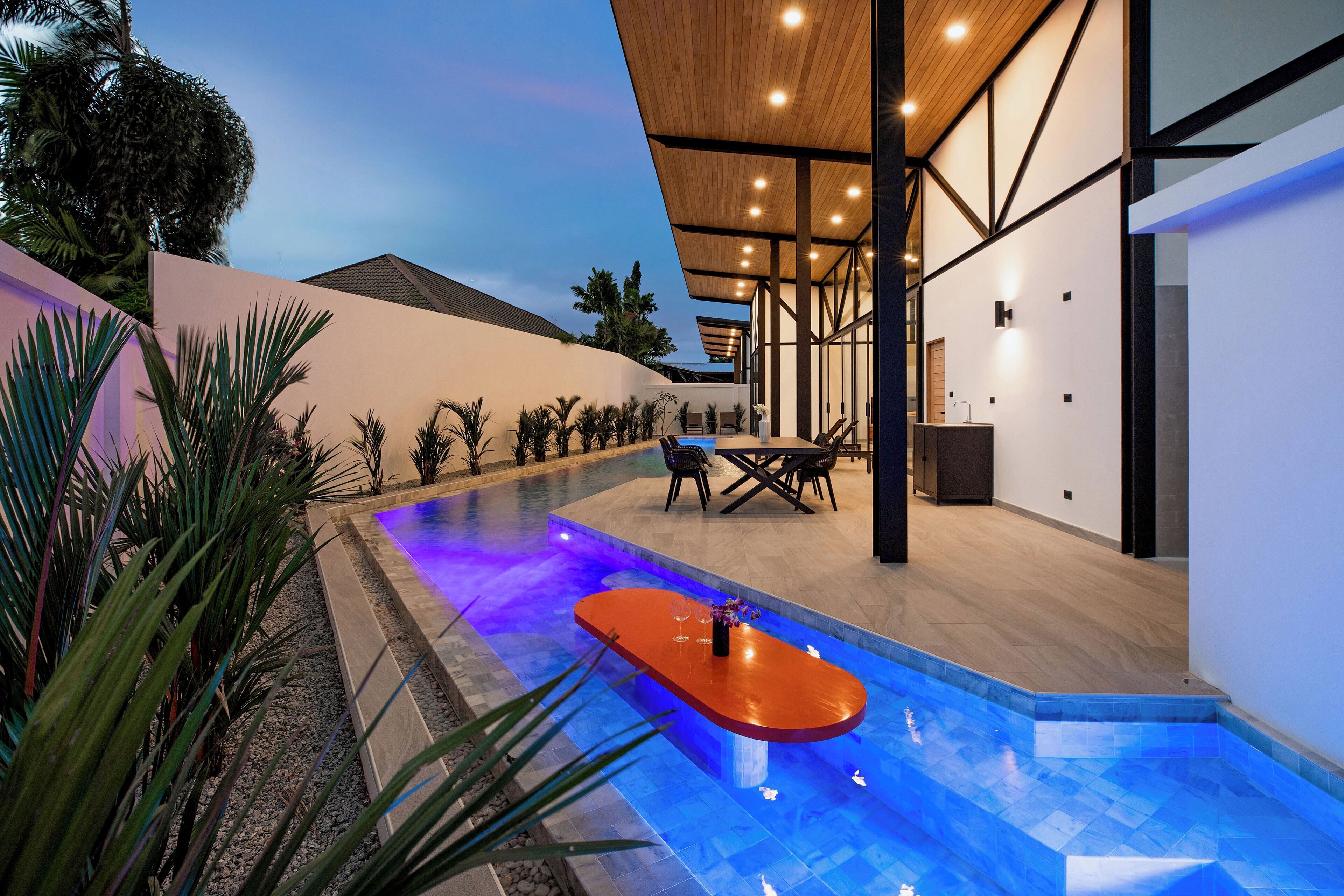 Uma área de piscina moderna ao ar livre com deck de madeira e assentos, rodeada por plantas tropicais.