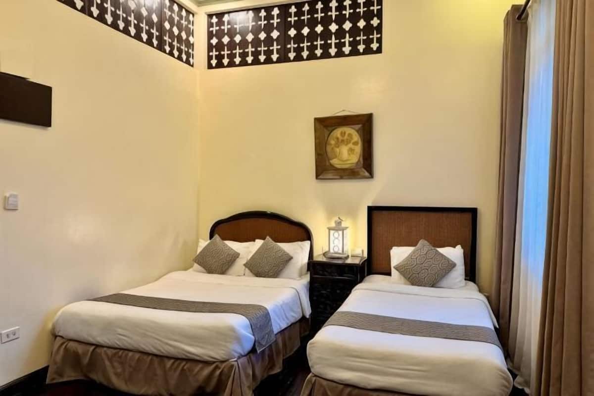 Habitación de hotel con dos camas, una mesita de noche con lámpara, un cuadro en la pared y una ventana con cortinas.