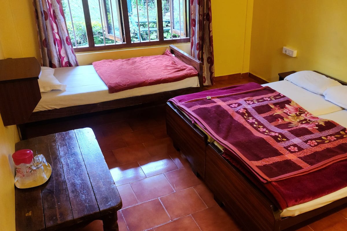 A room with two beds, one with a pink blanket and the other with a floral quilt. There is a small table with a bottle and a glass, and a window with curtains.