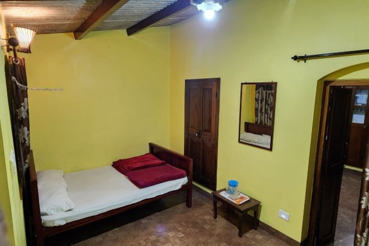 A small room with a single bed, a wooden door, a small table with a lamp, and a mirror on the wall.