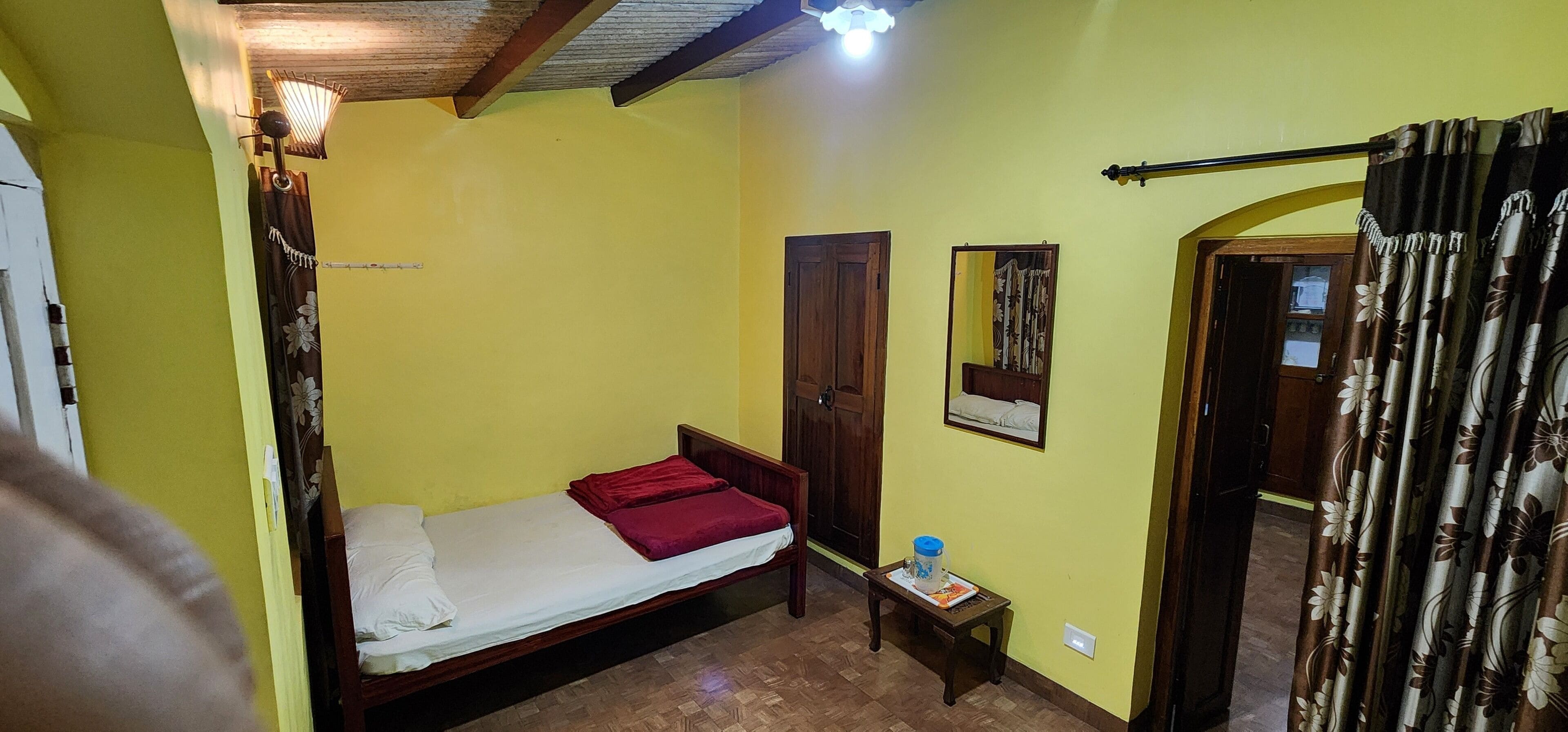 A small room with a single bed, a wooden door, a small table with a water bottle, and a mirror on the wall.