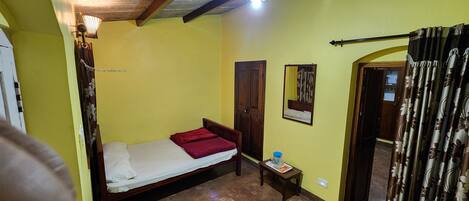 A small room with a single bed, a wooden door, a small table with a water bottle, and a mirror on the wall.