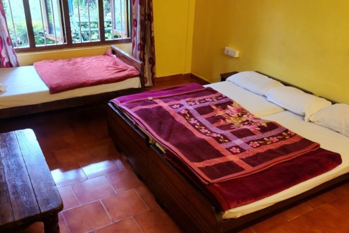 A room with two beds, one with a floral quilt and the other with a plain white bedspread. There is a wooden bench and a window with curtains.