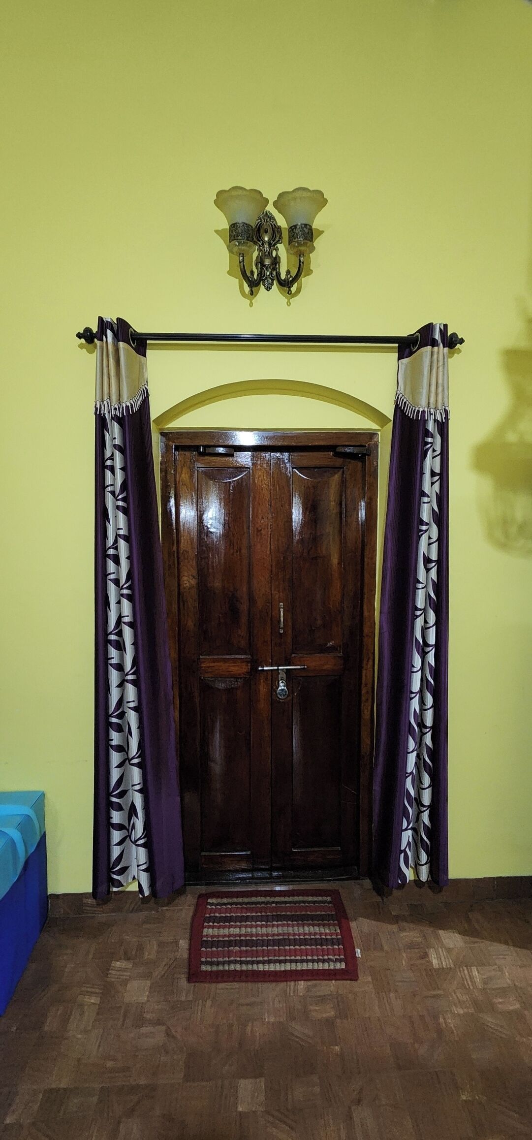 Una puerta de madera con un picaporte de metal, enmarcada por cortinas de tela morada y blanca, y una alfombra pequeña sobre un suelo de madera.