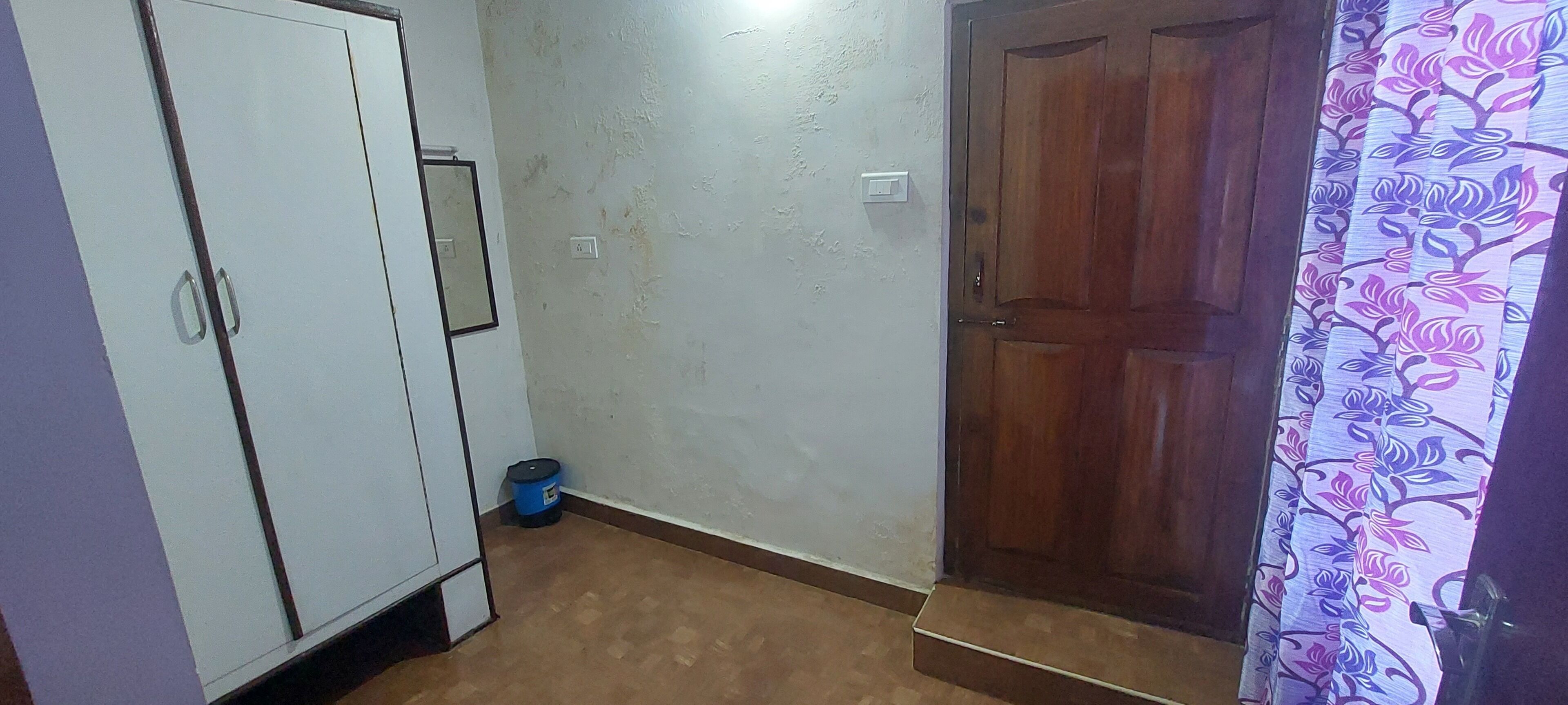 Ein Zimmer mit einem Schrank, einer Holztür, einem Mülleimer und einem gemusterten Vorhang.