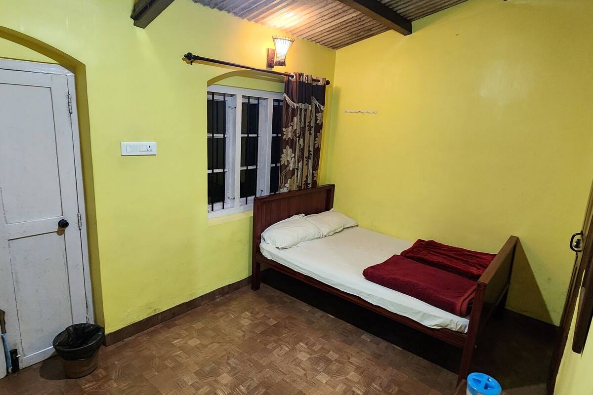 A small, single-bed room with a wooden bed, a window with curtains, a door, and a light fixture above the bed.