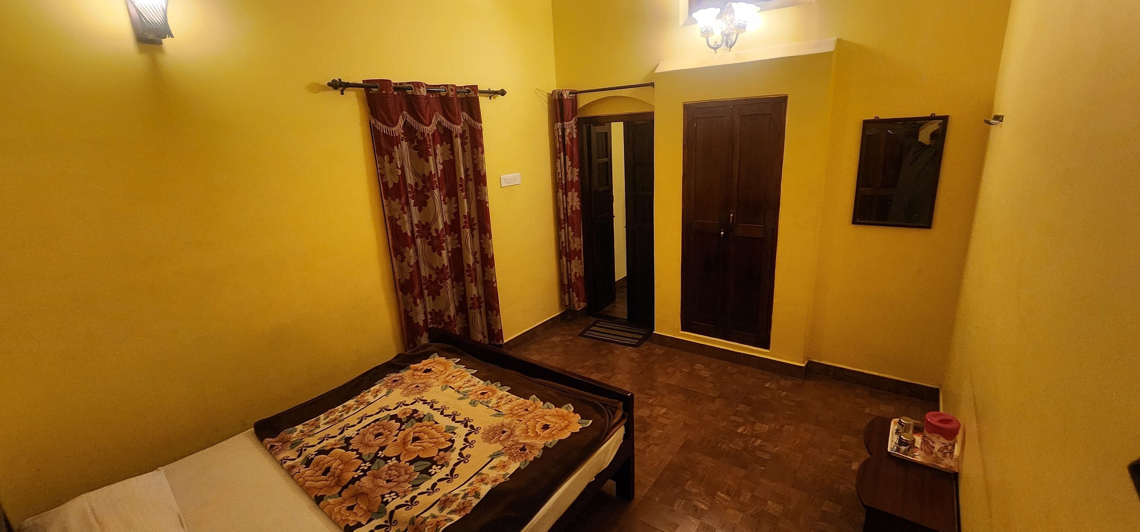 A room with a bed, a small table, a chair, a door, and a wall-mounted light fixture.