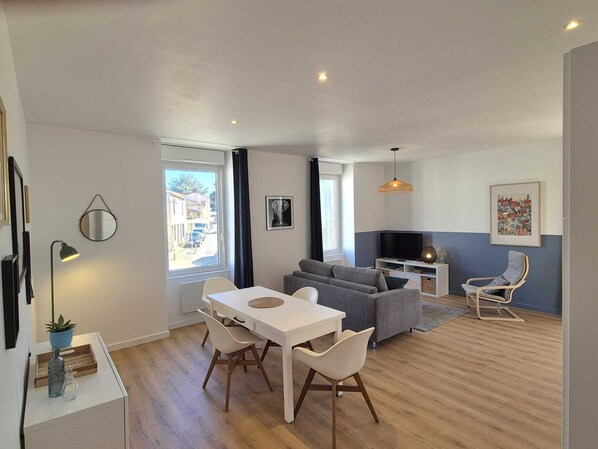 A modern living room with a sofa, dining table, and chairs. There is a TV and a small table with a lamp. - Apartment n°2: Marguerite (La Gaubretière)