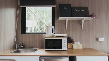 A compact kitchen with a microwave, coffee maker, and a small refrigerator. A window with a black roller blind is above the sink, and there are two shelves with a welcome sign and a box labeled "Escape".