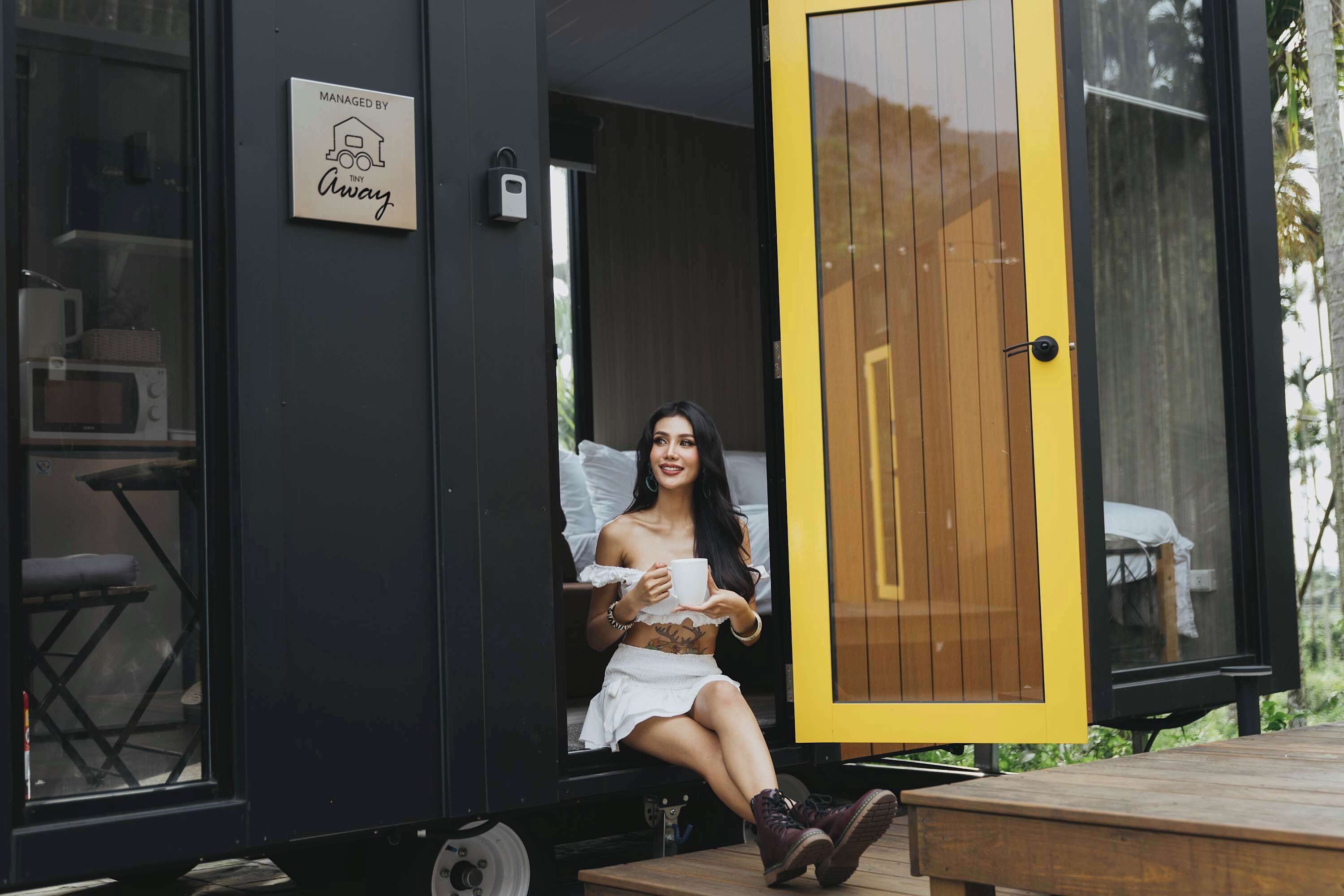 A person sitting on a bench outside a modern, compact living space with a wooden door and a sign that reads "MANAGED BY Tiny Away."