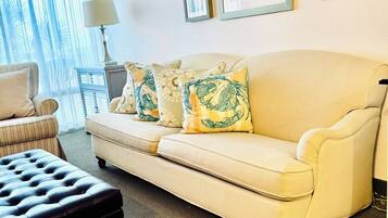 Ein Wohnzimmer mit einem beigefarbenen Sofa, einem schwarzen Ottomanen und einem Sessel. An den Wänden hängen gerahmte Bilder und auf einem Beistelltisch steht eine Lampe.