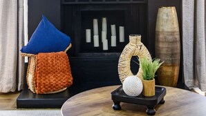 Un salon moderne avec une cheminée sombre, une étagère en bois ornée d’objets décoratifs, un coussin bleu et une table ronde en bois sur laquelle reposent un panier tressé et une plante.