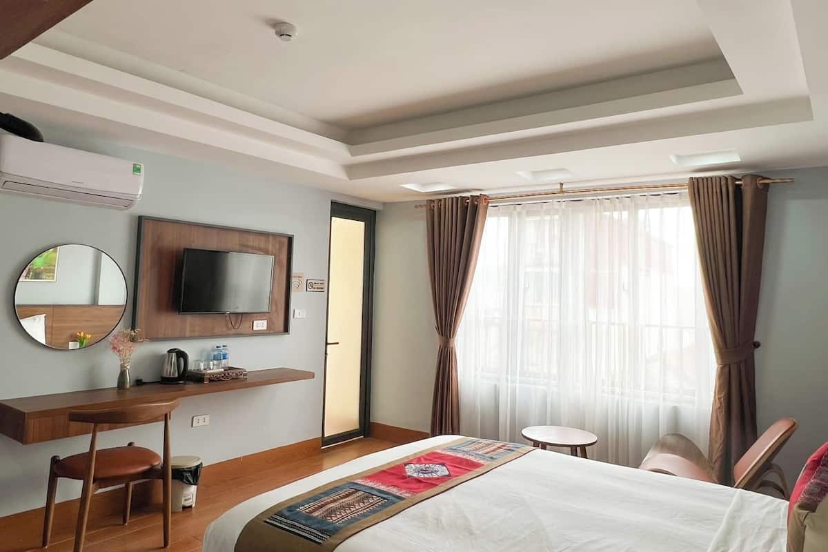 Een hotelkamer met een bed, een bureau met stoel, een televisie, een spiegel en een raam met gordijnen.