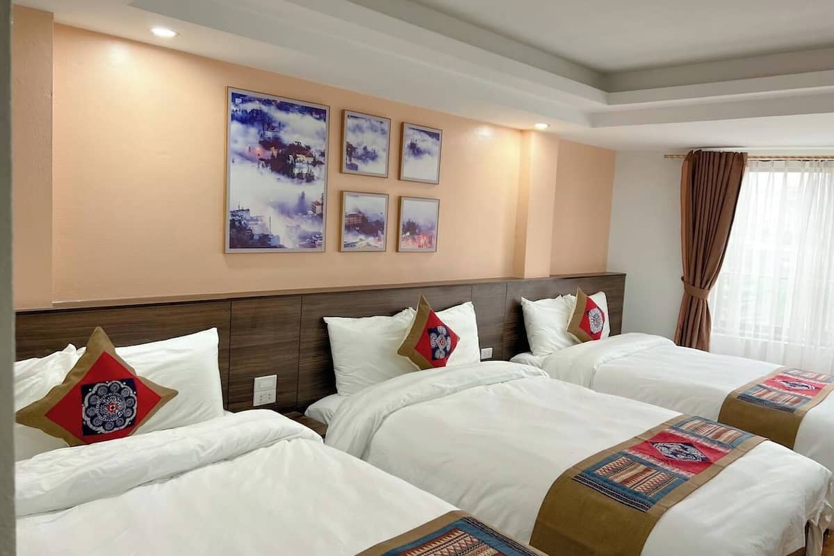 Hotelkamer met vier bedden op rij, elk voorzien van witte beddengoed en decoratieve kussens. De kamer heeft een houten hoofdbord, een groot raam met gordijnen en wanddecoraties met landschappen.