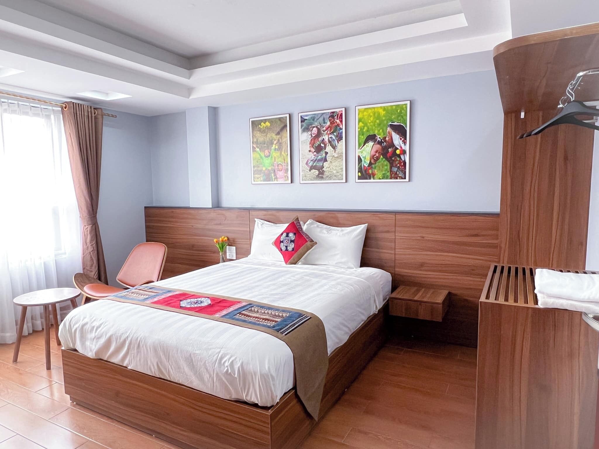 A hotel room with a wooden bed, a small table, a chair, and a wall with three framed pictures.