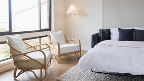A bedroom with a large bed, two wicker chairs, a floor lamp, and a view through a window. - Ocean View Escape, Gerroa (Gerroa)
