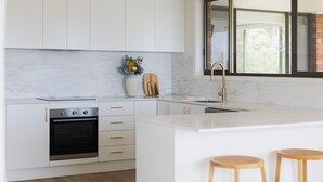 A modern kitchen with white cabinetry, a built-in oven, a large island with stools, and a window with a view. - Ocean View Escape, Gerroa (Gerroa)