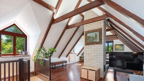 A spacious attic room with wooden beams, a brick fireplace, a television, and a window with a view of greenery.