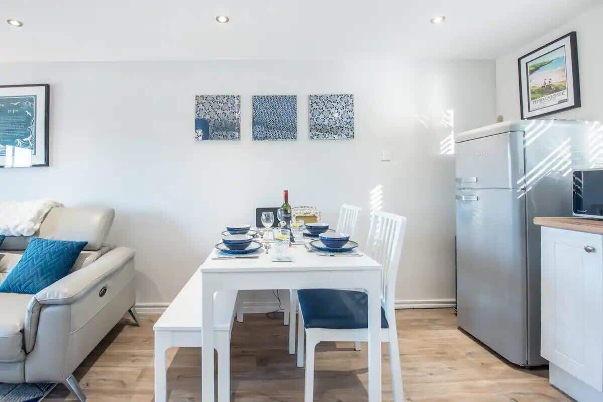 Una sala moderna con un sofá blanco, una mesa de comedor con platos azules, y una zona de cocina con refrigerador y microondas.