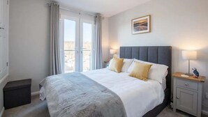 A hotel room with a large bed, a bedside table with a lamp, a small bench, and a window with curtains. - Sea View Chalet | Walk to Freshwater Beach (Pembrokeshire)