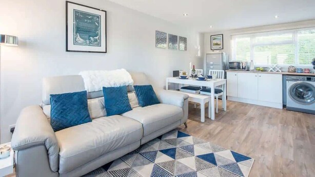 A modern living room with a sofa, a dining table, and a kitchen area including a washing machine. - Sea View Chalet | Walk to Freshwater Beach (Pembrokeshire)