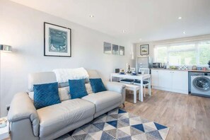 A modern living room with a sofa, a dining table, and a kitchen area including a washing machine. - Sea View Chalet | Walk to Freshwater Beach (Pembrokeshire)
