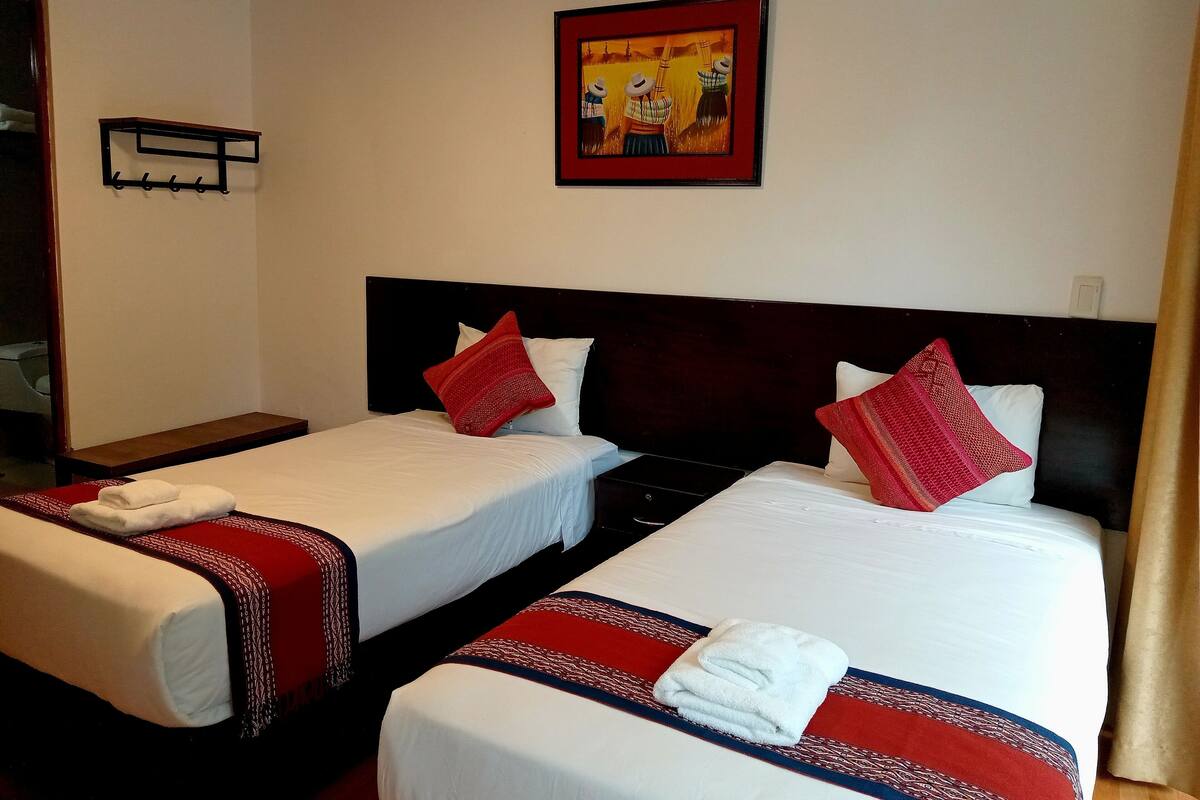 A hotel room with two single beds, each with a red and white patterned bedspread, white pillows, and a white towel folded on the bed. There is a framed picture on the wall above the beds.