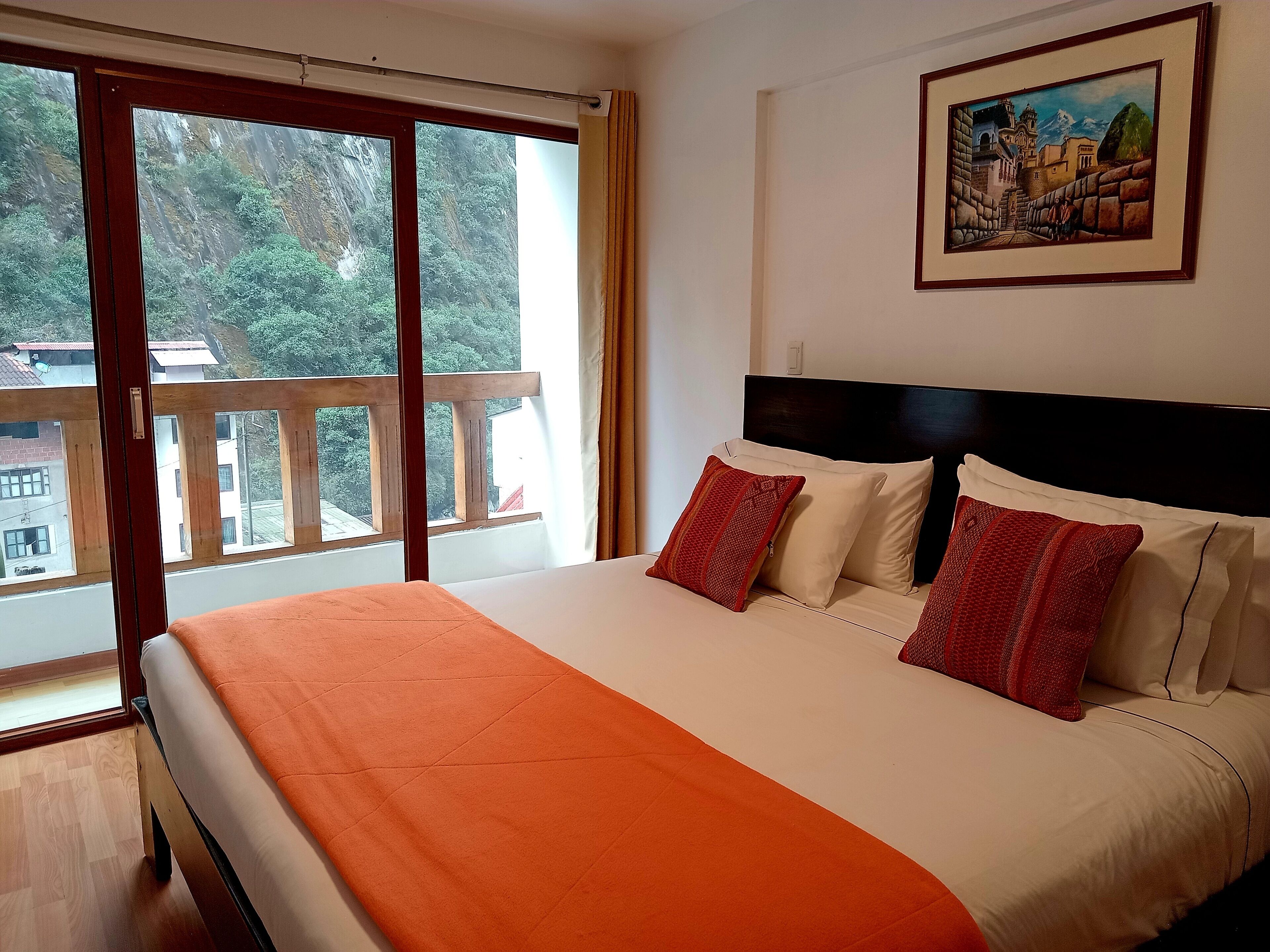 A hotel room with a large bed, a painting on the wall, and a balcony with a view of trees and buildings.