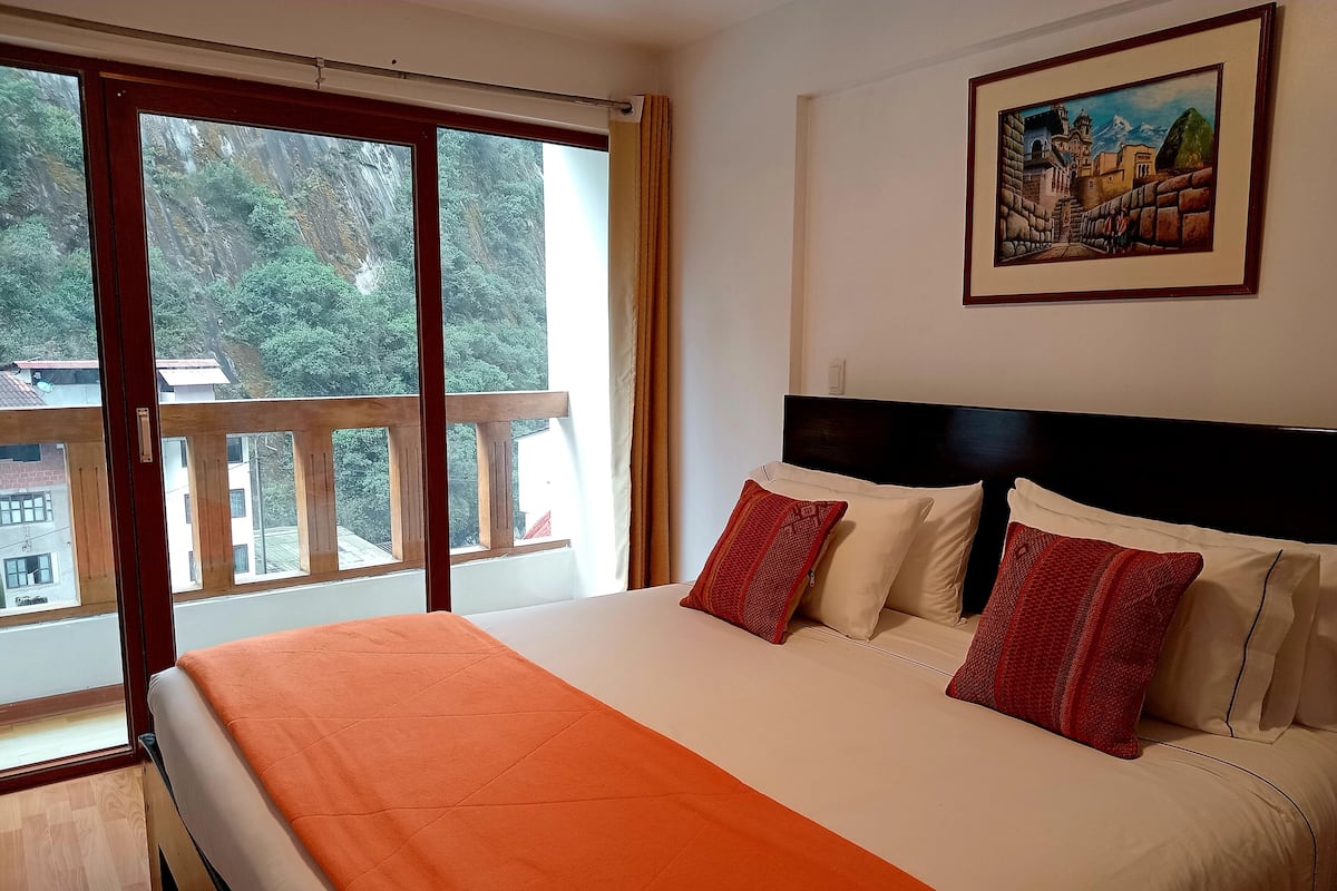 A hotel room with a large bed, a painting on the wall, and a balcony with a view of trees and buildings.