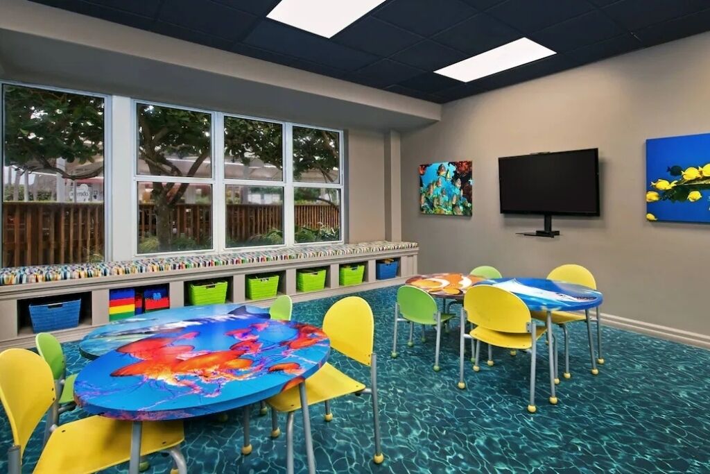 Una sala de juegos para niños con mesas y sillas coloridas, un ventanal con vistas a árboles y obras de arte en las paredes.